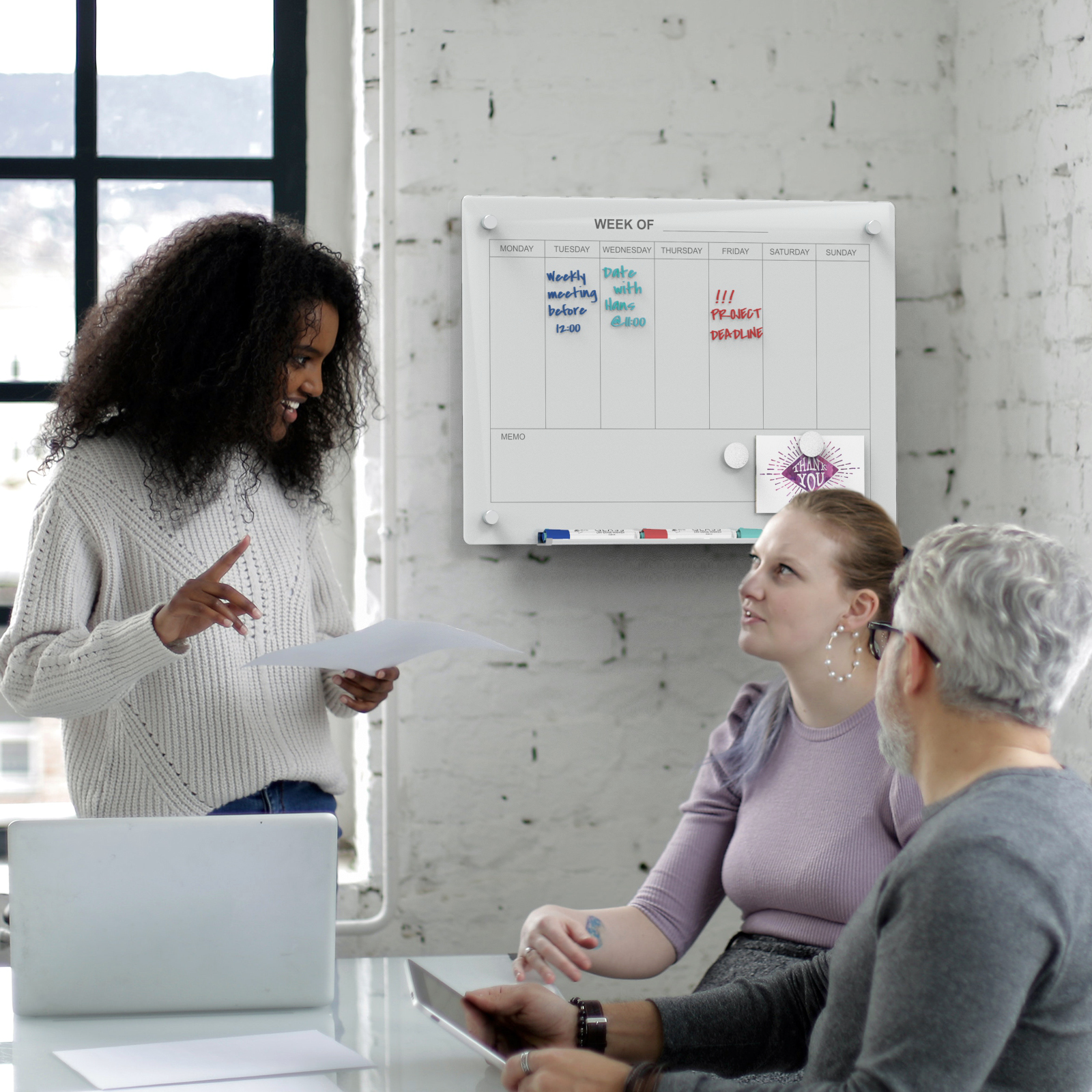 Wall mounted weekly planner board with group discussing projects in front of the board. 