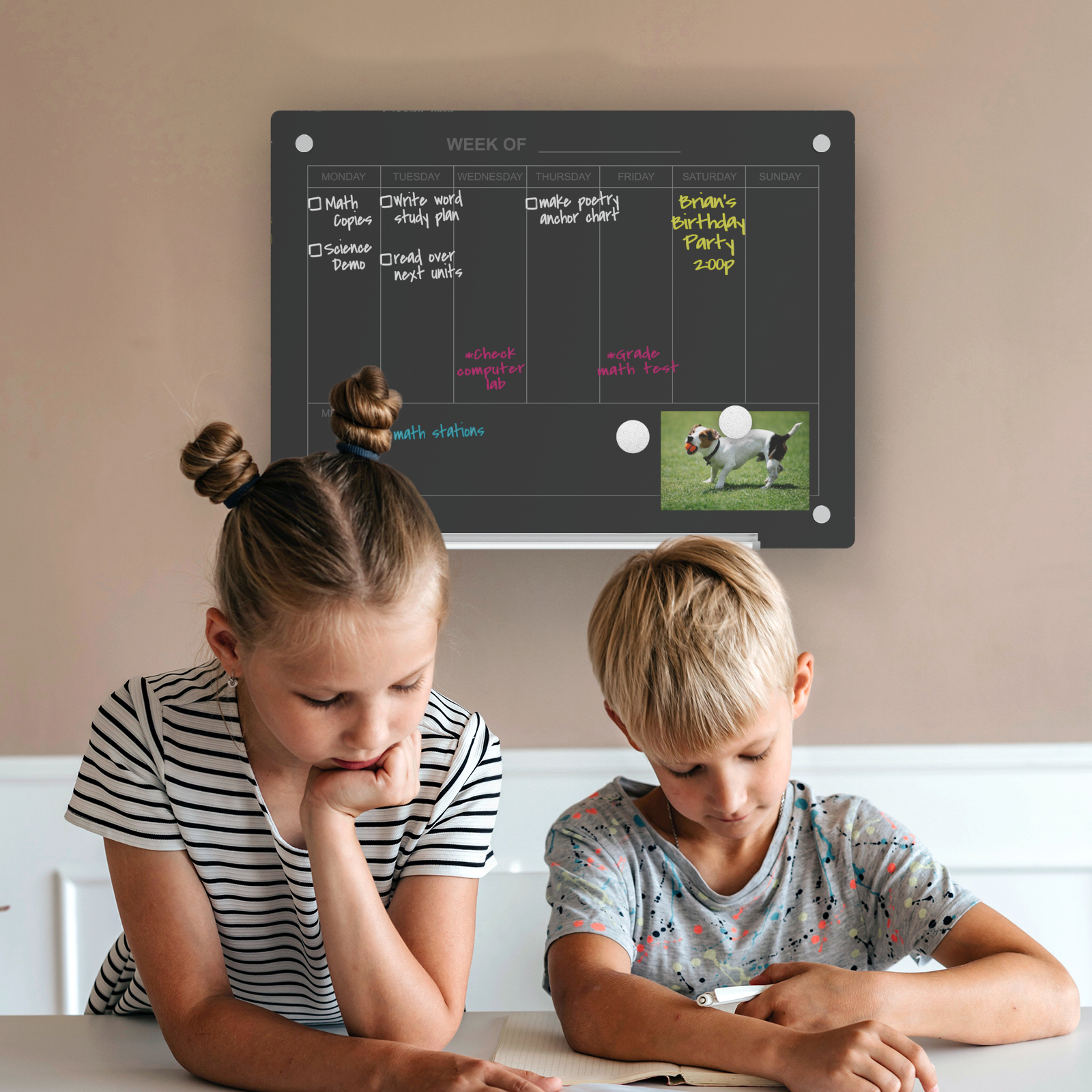 Wall mounted black dry erase planner behind two kids studying. 