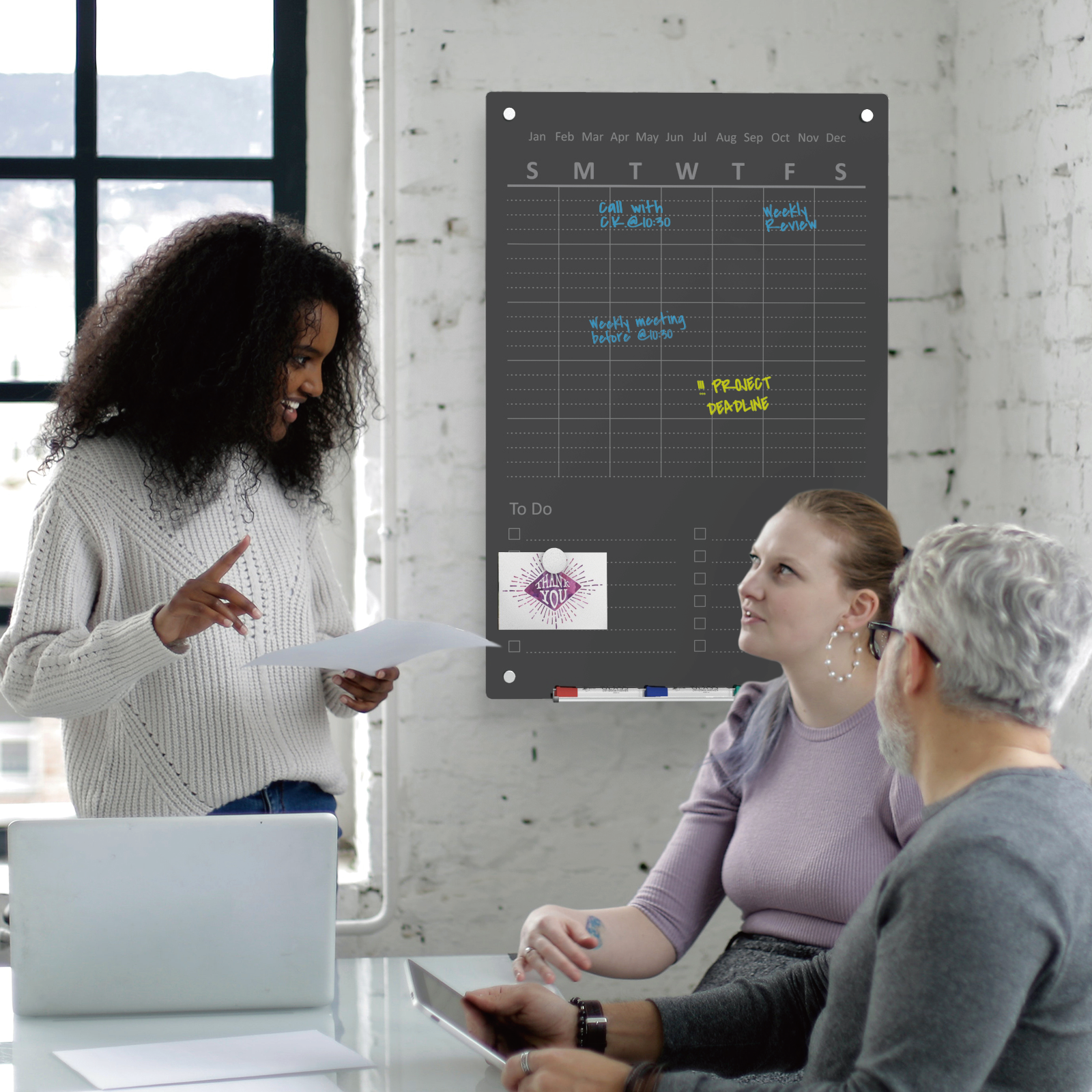 Audio-Visual Direct Glass Wall Mounted Chore Board / To Do list planner weekly in black.  Lifestyle image showing a group discussing it in an office setting. 